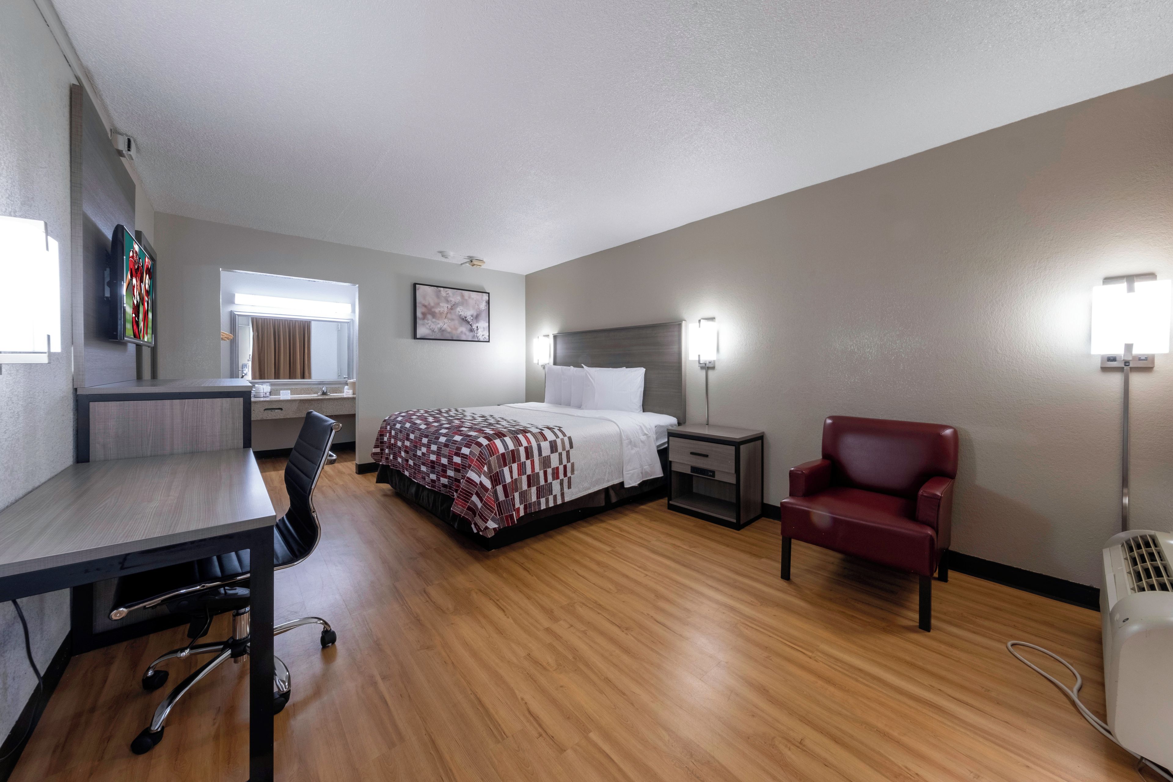 A hotel room with a bed, bedside table, chair, desk, and a TV.