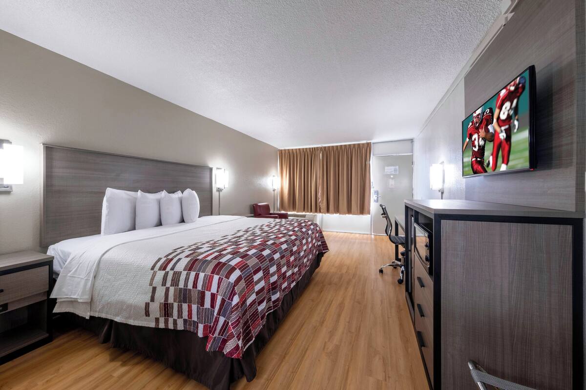 A hotel room with a large bed, a desk, a TV mounted on the wall, and a window with curtains.