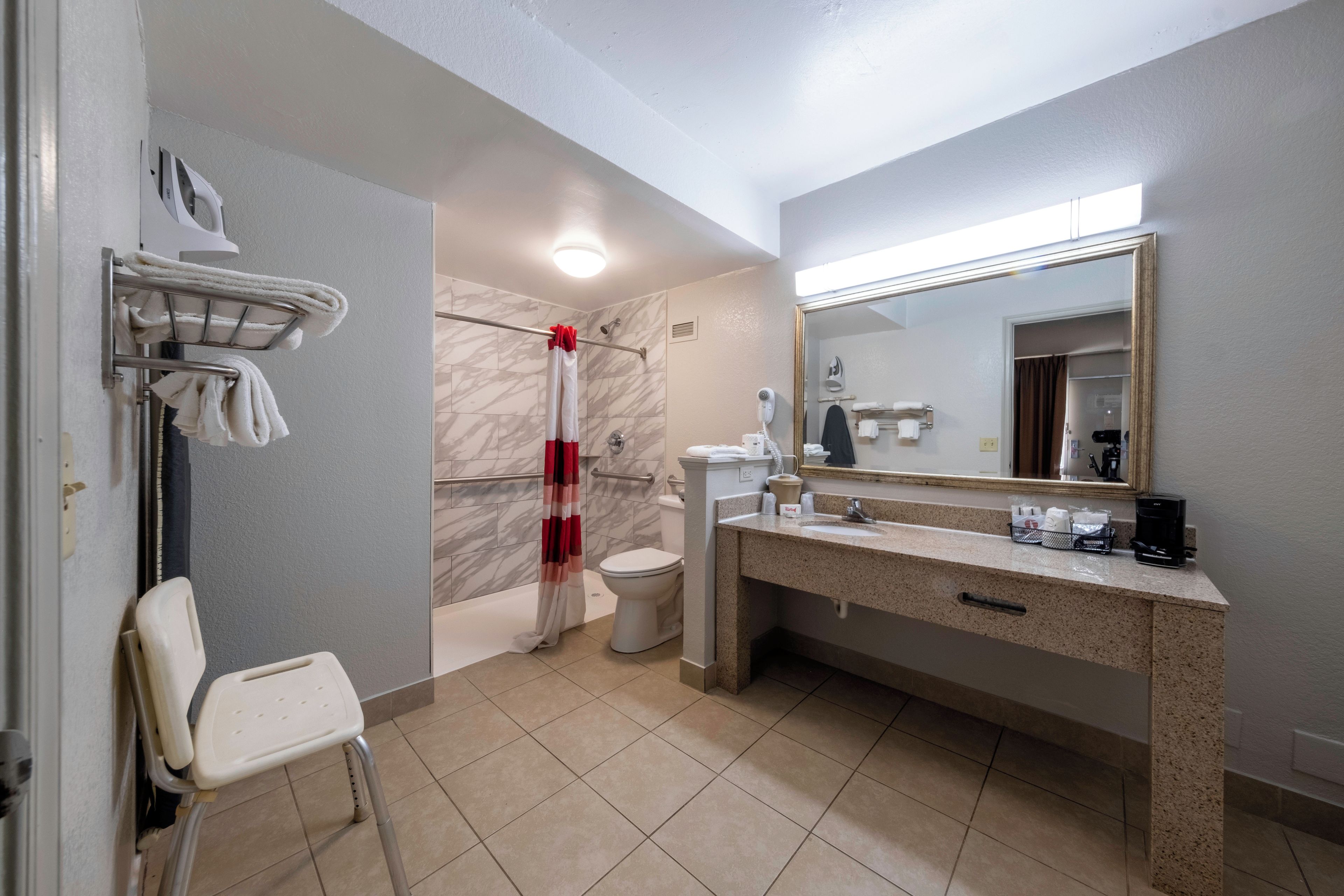 A bathroom with a shower, toilet, and sink. There is a towel rack with towels, a chair, and a mirror with a vanity.