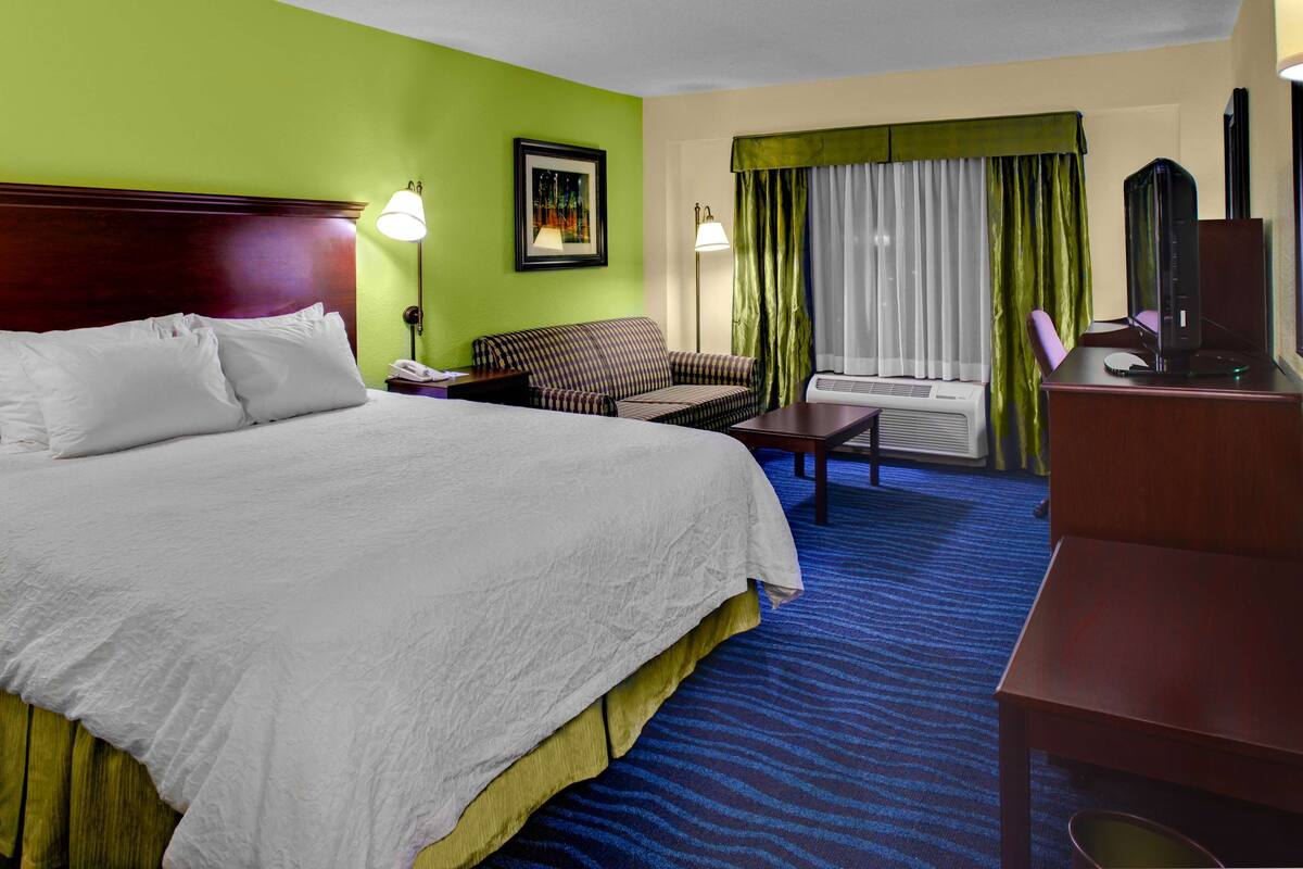 A hotel room with a large bed, a sofa, a desk with a computer, and a TV.