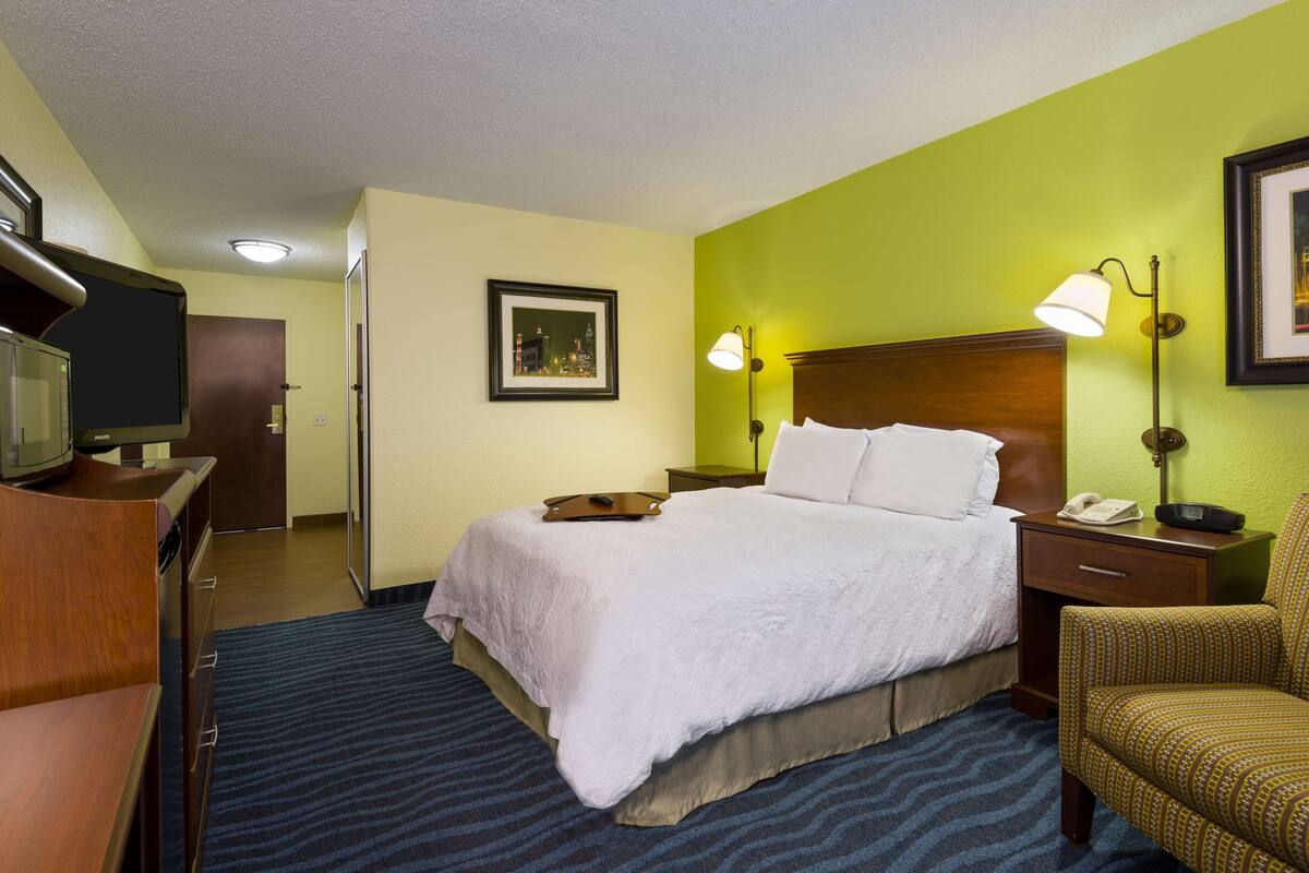 A hotel room with a bed, bedside lamps, a chair, a desk with a television, and a framed picture on the wall.