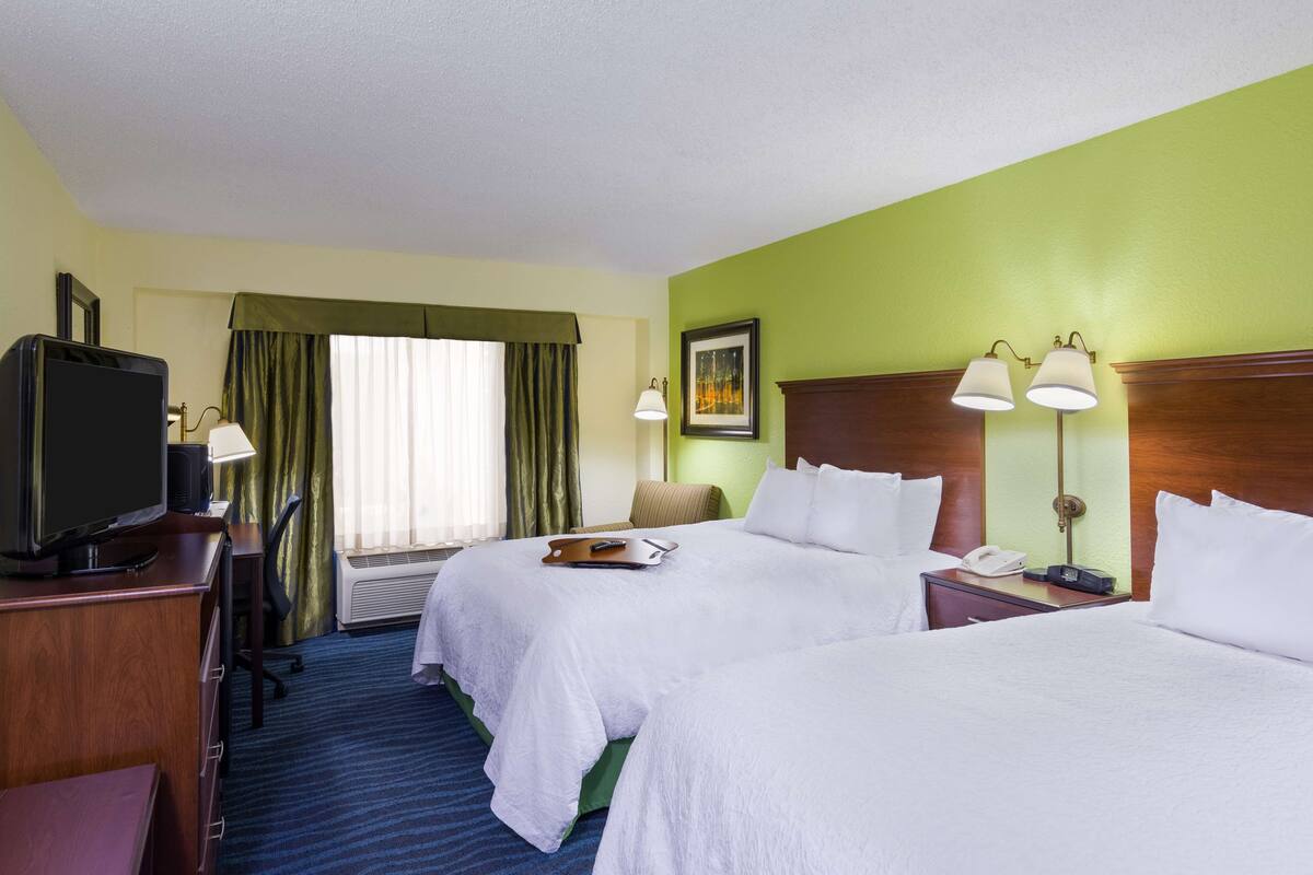 A hotel room with two beds, a desk with a television, a lamp, and a framed picture on the wall.