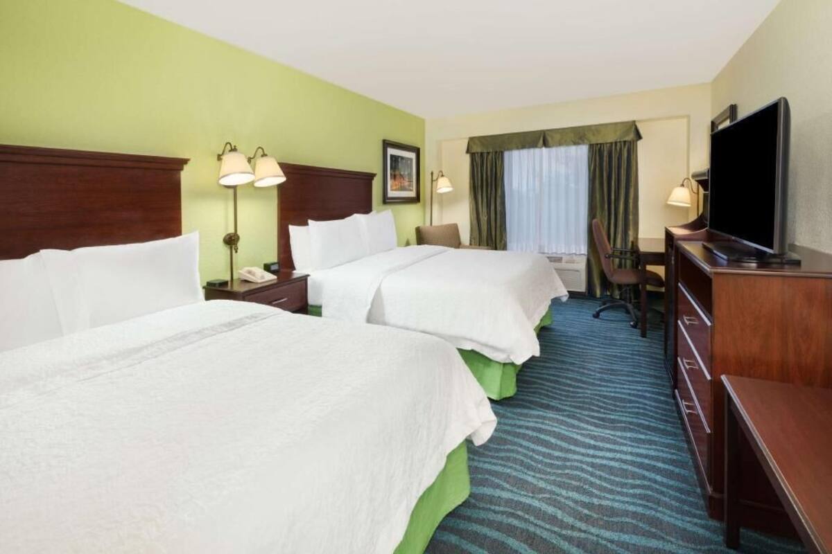 A hotel room with two beds, a desk, a television, and a window with curtains.