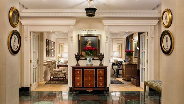 A grand hallway with marble flooring, a central wooden console, framed artwork, and a chandelier. - Hôtel Francois 1er (Paris)