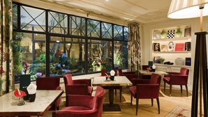A restaurant with red chairs and tables, a large window with a view of greenery, and a shelf with decorative items. - Hôtel Francois 1er (Paris)