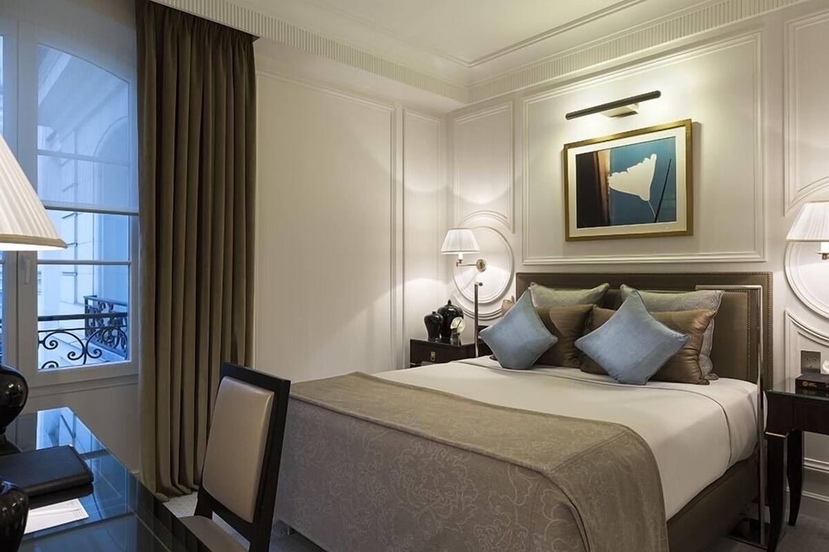 Ein Hotelzimmer mit einem großen Bett, einem Schreibtisch, einem Sessel, einem Fenster mit Vorhängen, einem Bild an der Wand und einer Lampe auf dem Nachttisch.