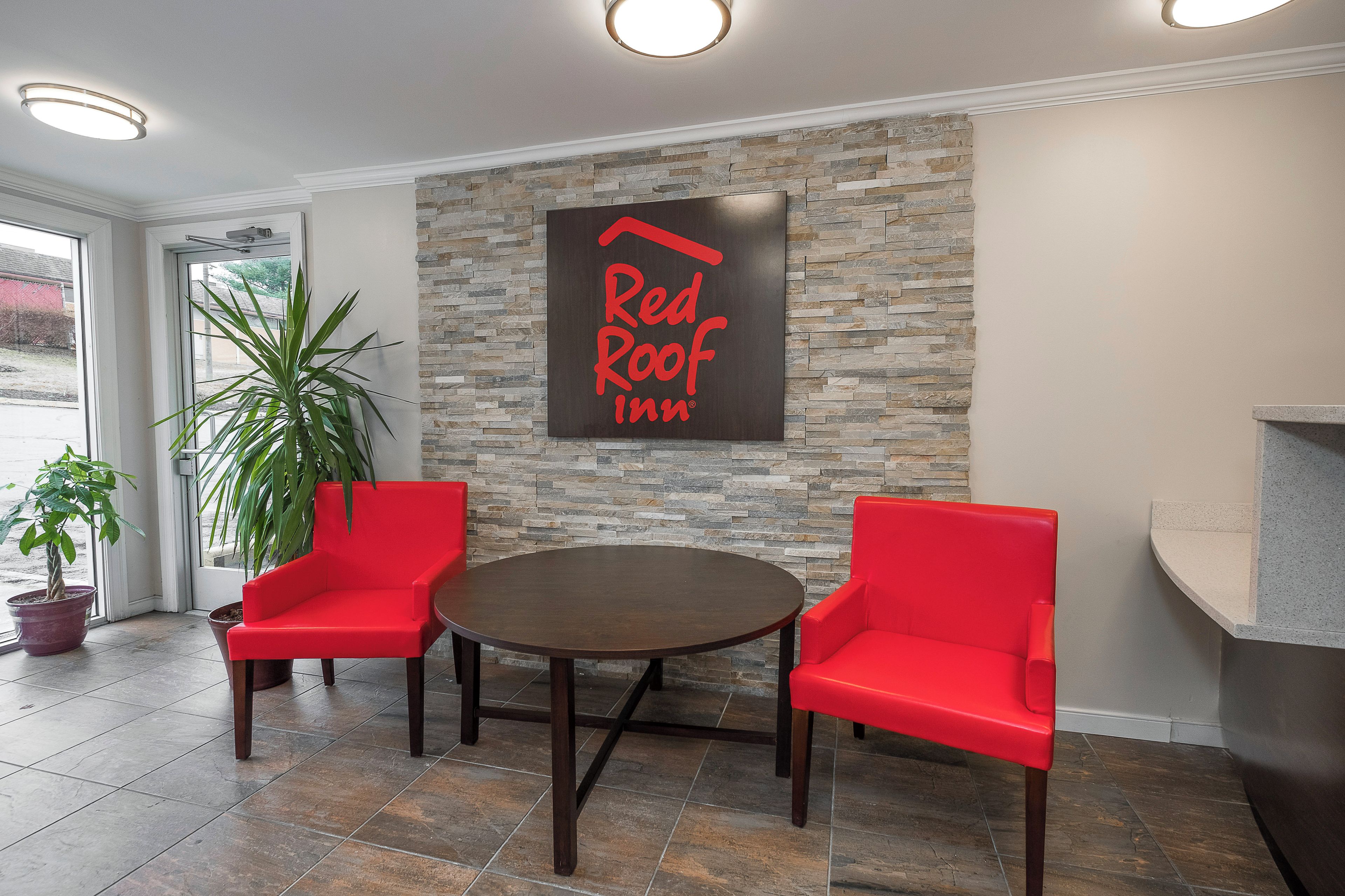 A Red Roof Inn reception area with a stone wall, two red chairs, a round table, and a potted plant.