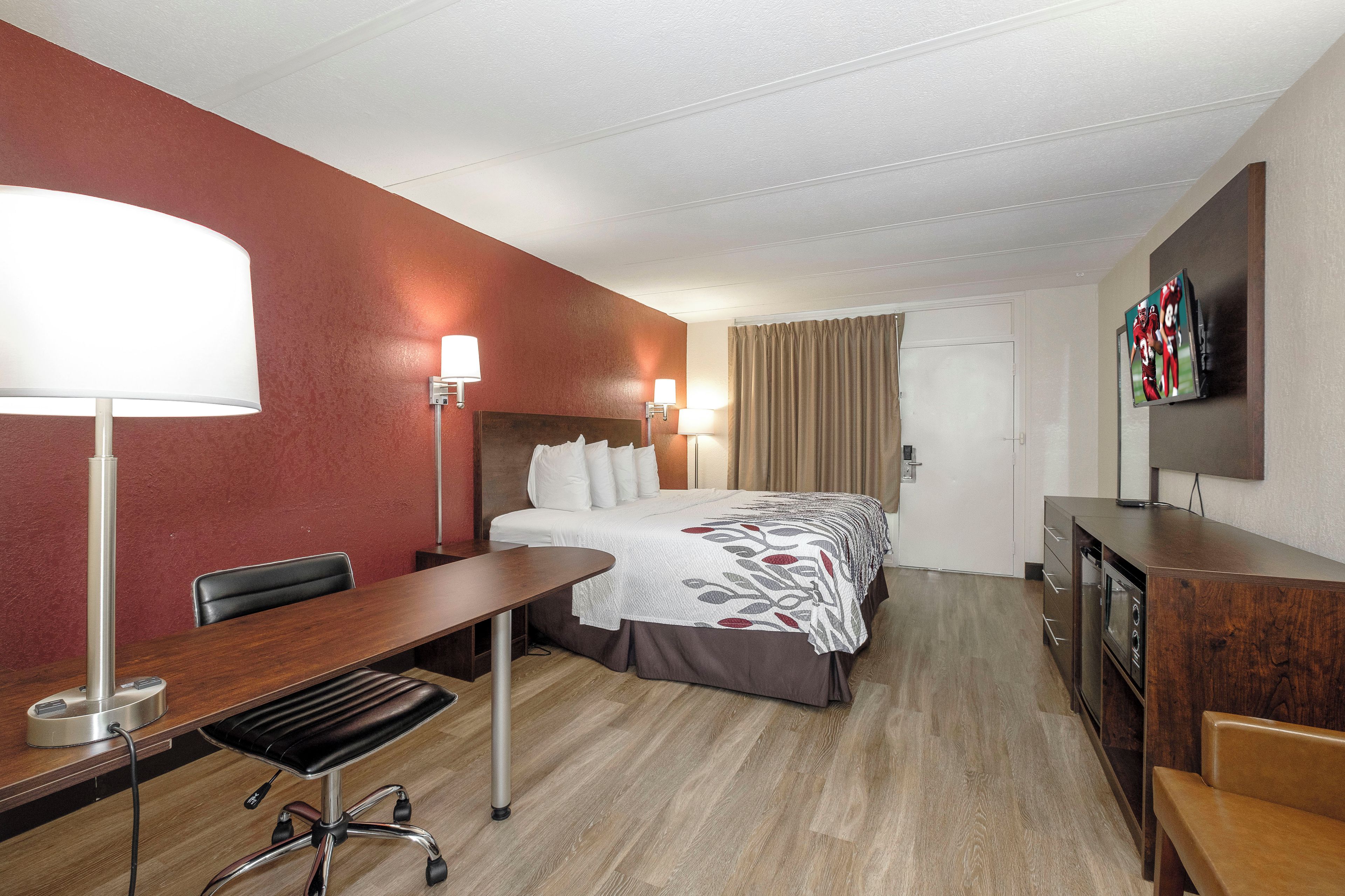 A hotel room with a bed, desk, chair, lamp, television, and a door.