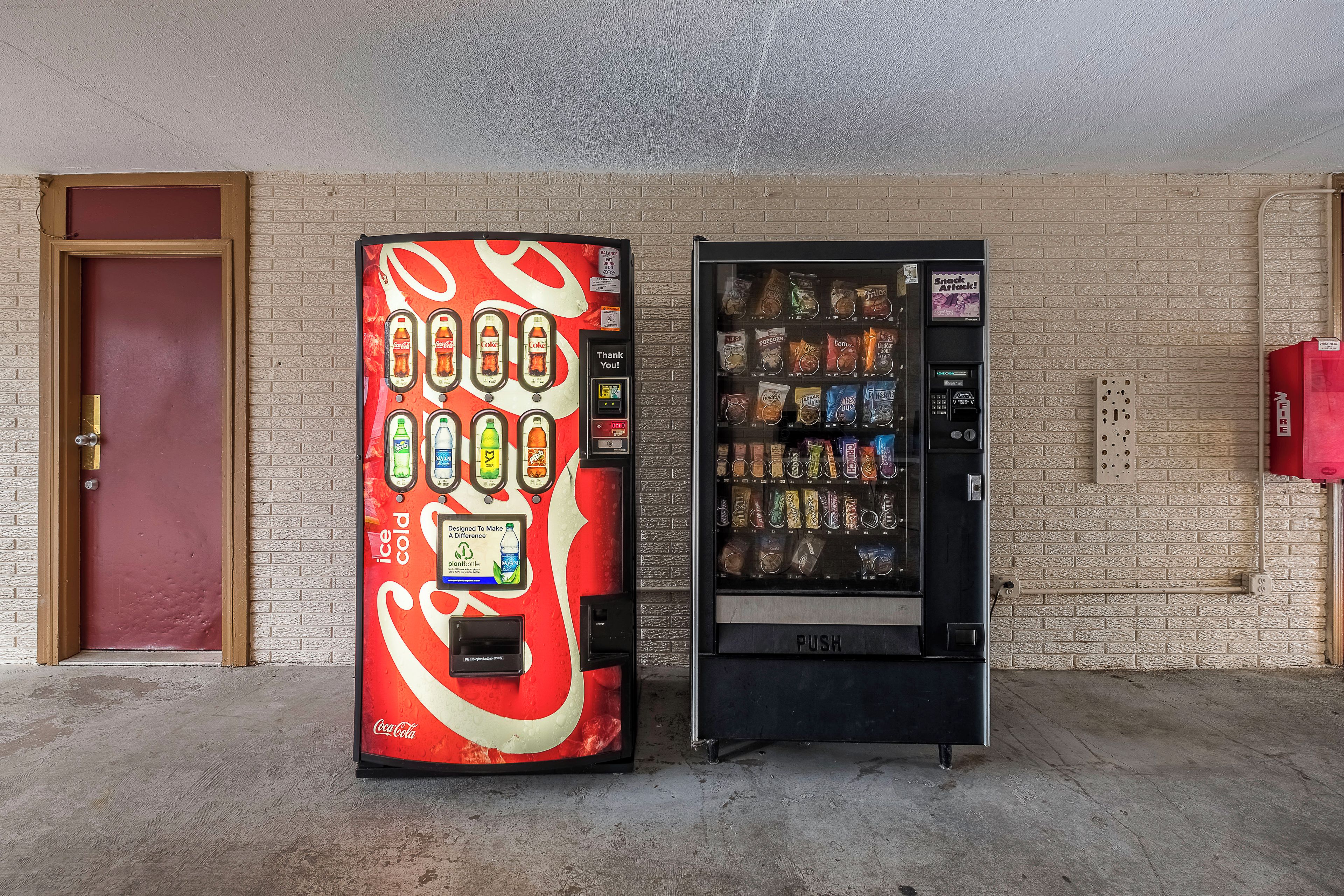 Dos máquinas expendedoras, una con el diseño de Coca-Cola y la otra con aperitivos, frente a un muro de ladrillo.