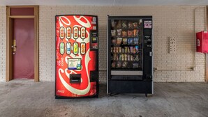 Twee automaten, waarvan één met een Coca-Cola ontwerp en de andere met snacks, tegen een bakstenen muur.