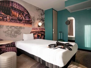 A hotel room with a bed, a seating area, a wall mural, a mirror, and a suitcase. - ibis styles Paris Montmartre Batignolles (Paris)