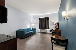 A modern living room with a sofa, a wooden table, a TV, and a wall-mounted lamp. - HomeTowne Studios by Red Roof East Lansing - Okemos (Okemos)
