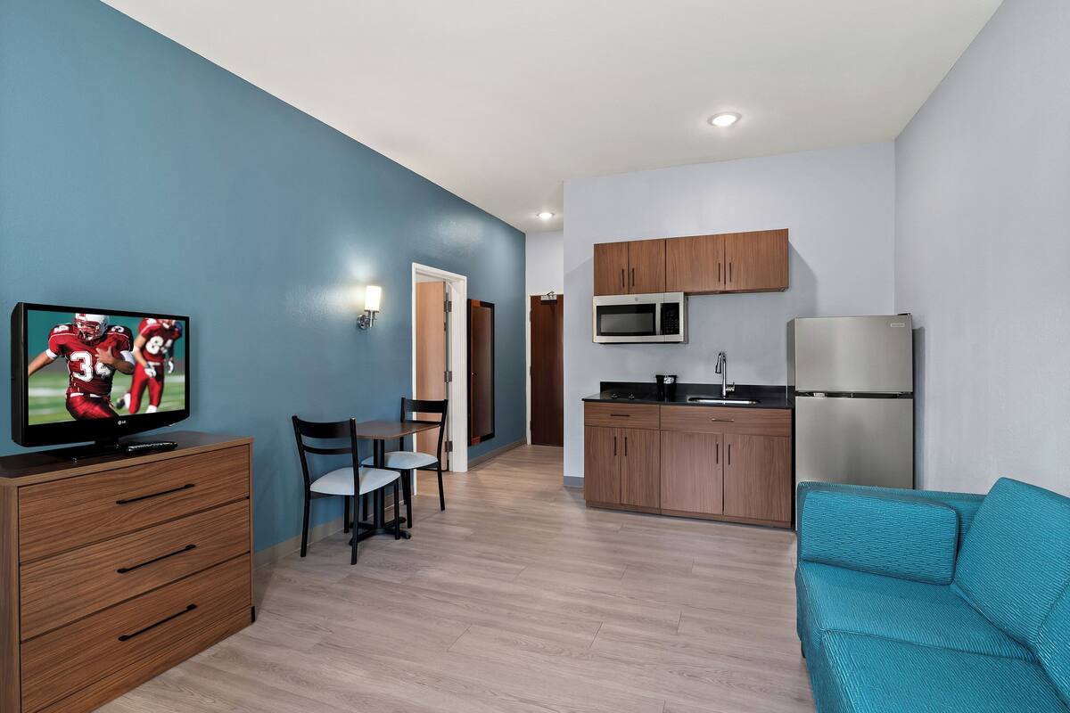 A modern living room with a flat-screen TV, a teal sofa, and a dining area with a table and chairs.
