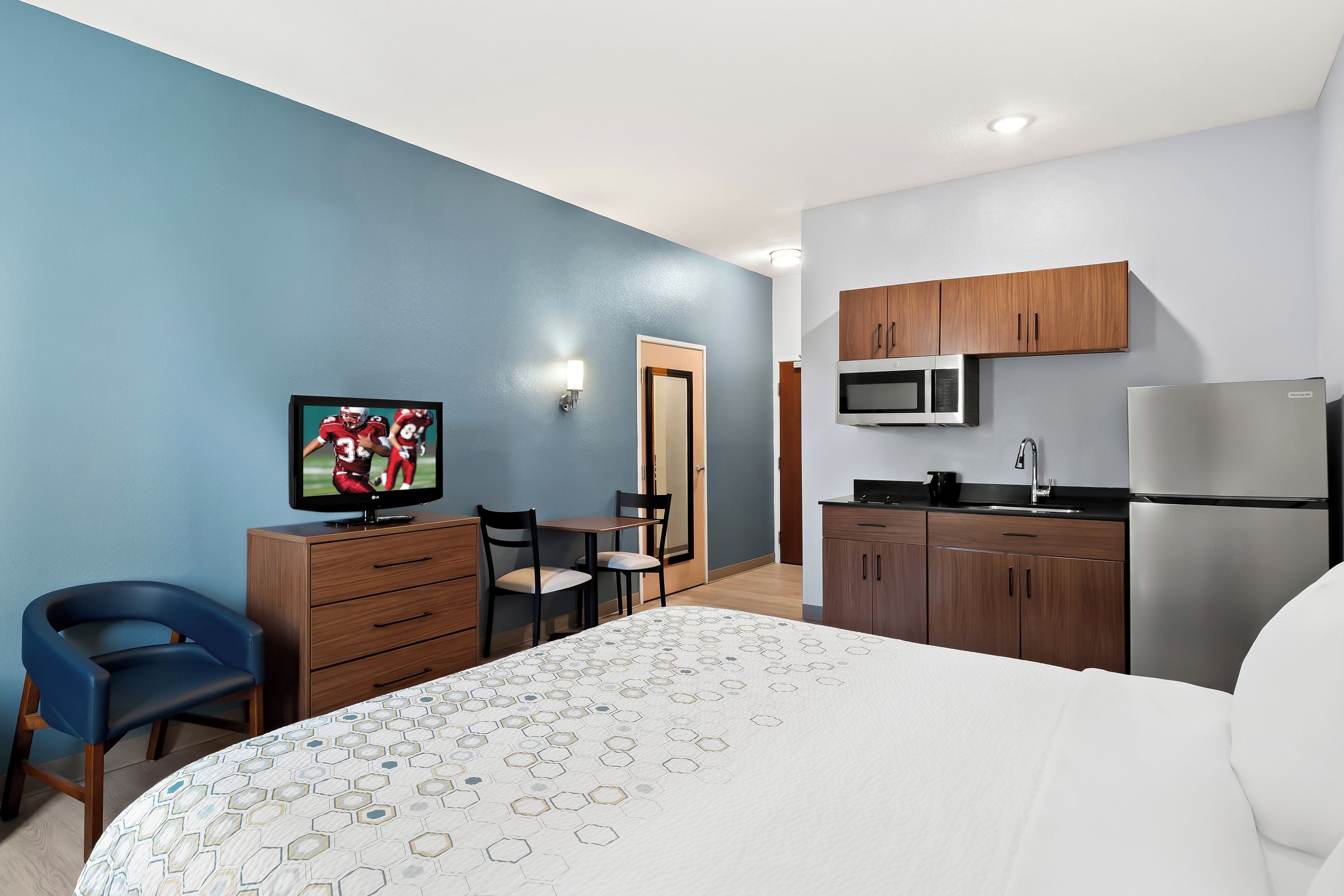A hotel room with a bed, a TV, a desk, chairs, a microwave, a refrigerator, and wooden cabinets.