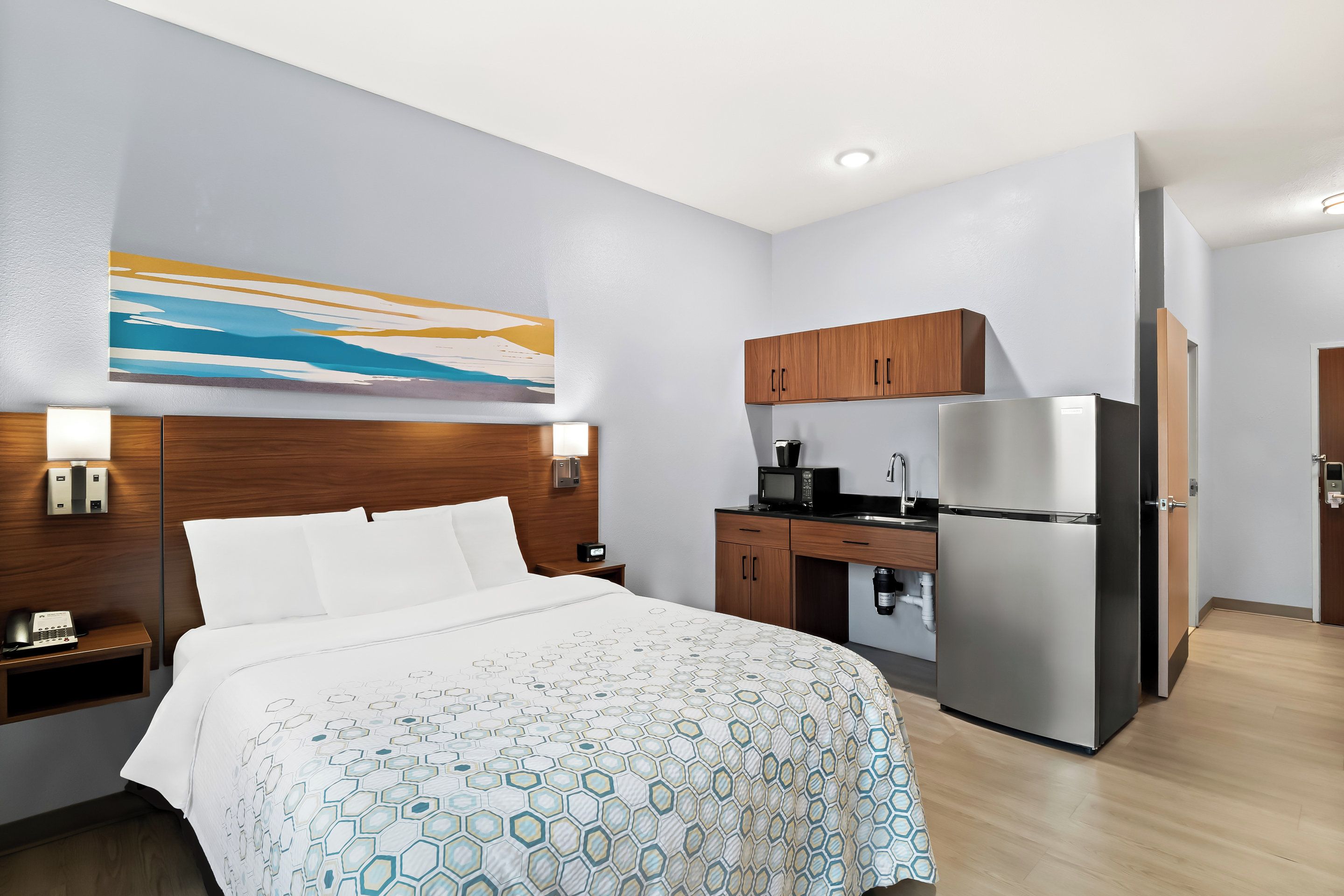 A hotel room with a bed, a nightstand, a desk, a refrigerator, and a kitchen area.