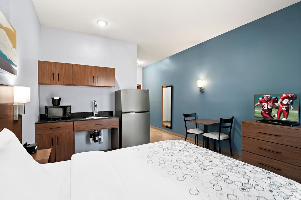 A modern hotel room with a kitchenette, a bed with a geometric-patterned bedspread, a flat-screen TV, and a large mirror.