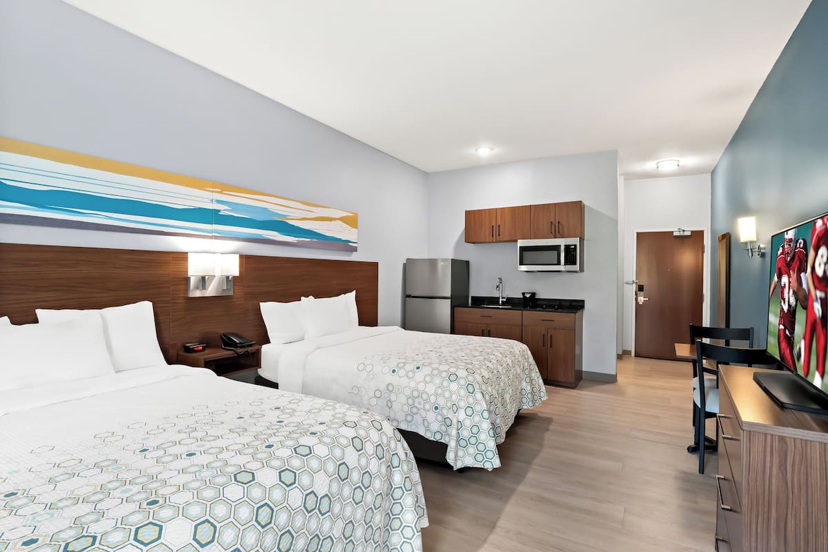 A hotel room with two beds, a kitchenette, a TV, and a painting on the wall.