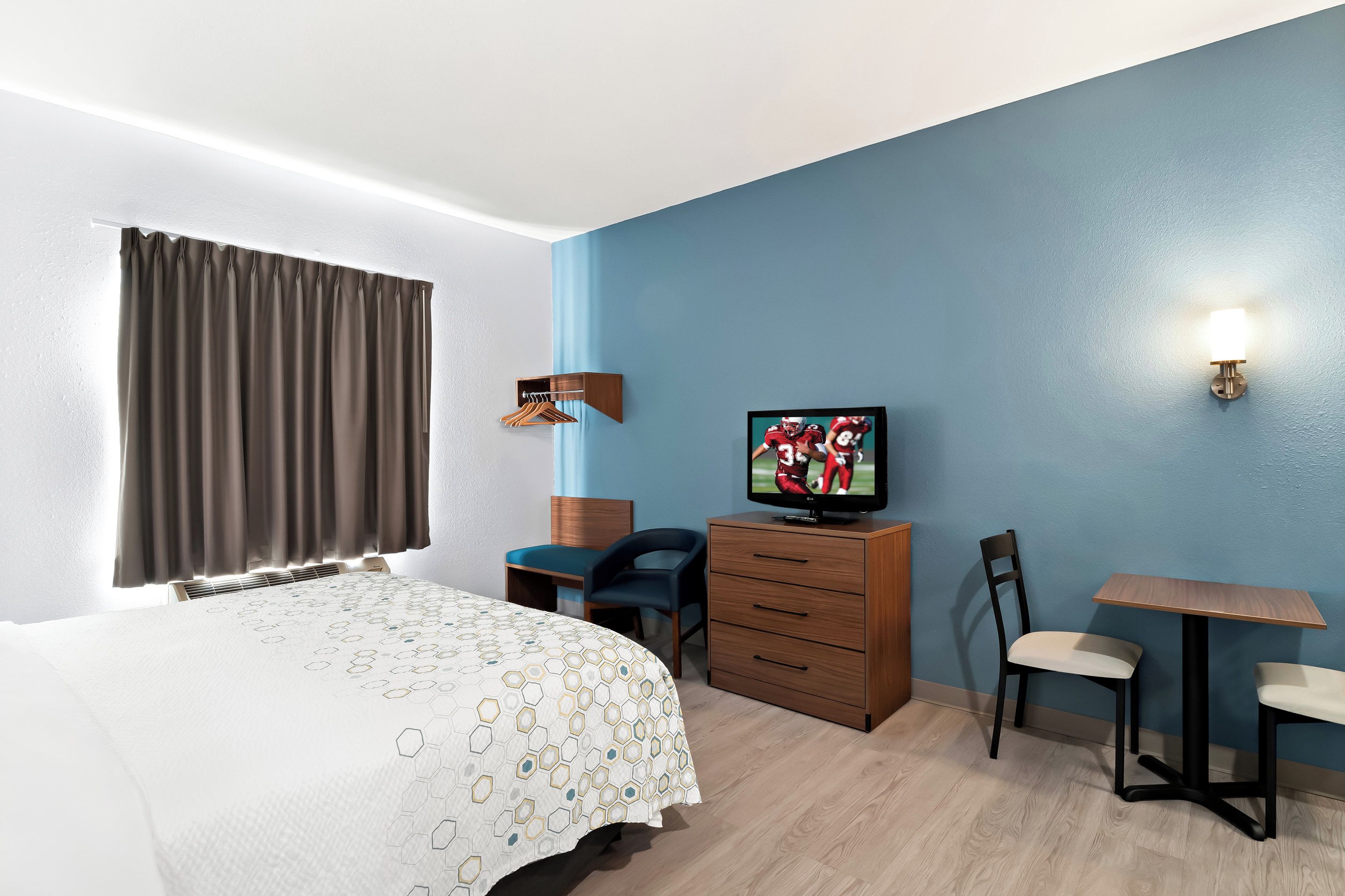 Quarto de hotel com cama, escrivaninha, cadeira, televisão e luminária na parede.