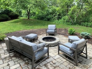 An outdoor seating area with a fire pit, surrounded by stone tiles and greenery.