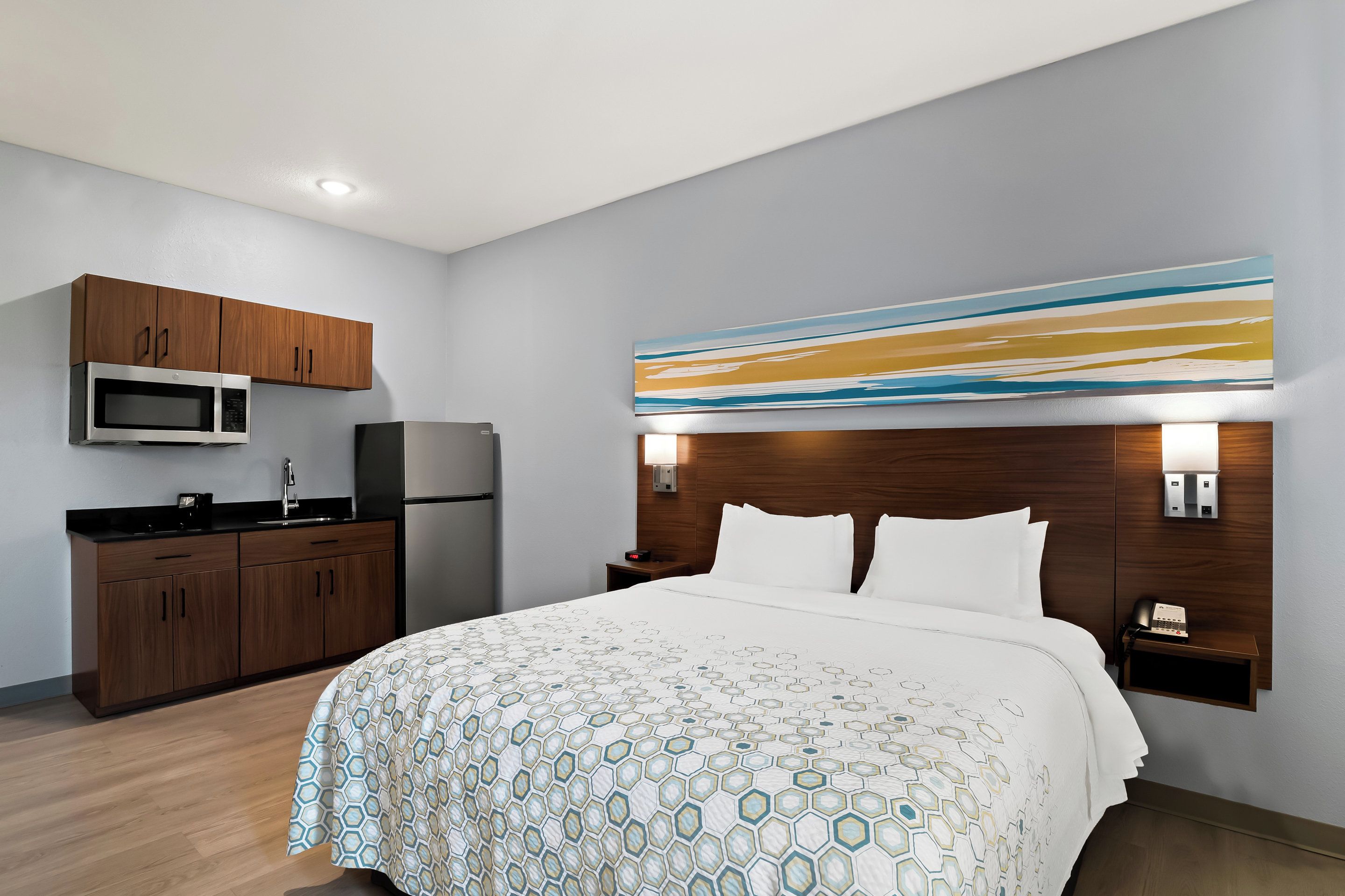 A hotel room with a bed, a wooden cabinet with a microwave, a refrigerator, and a small table with a lamp.