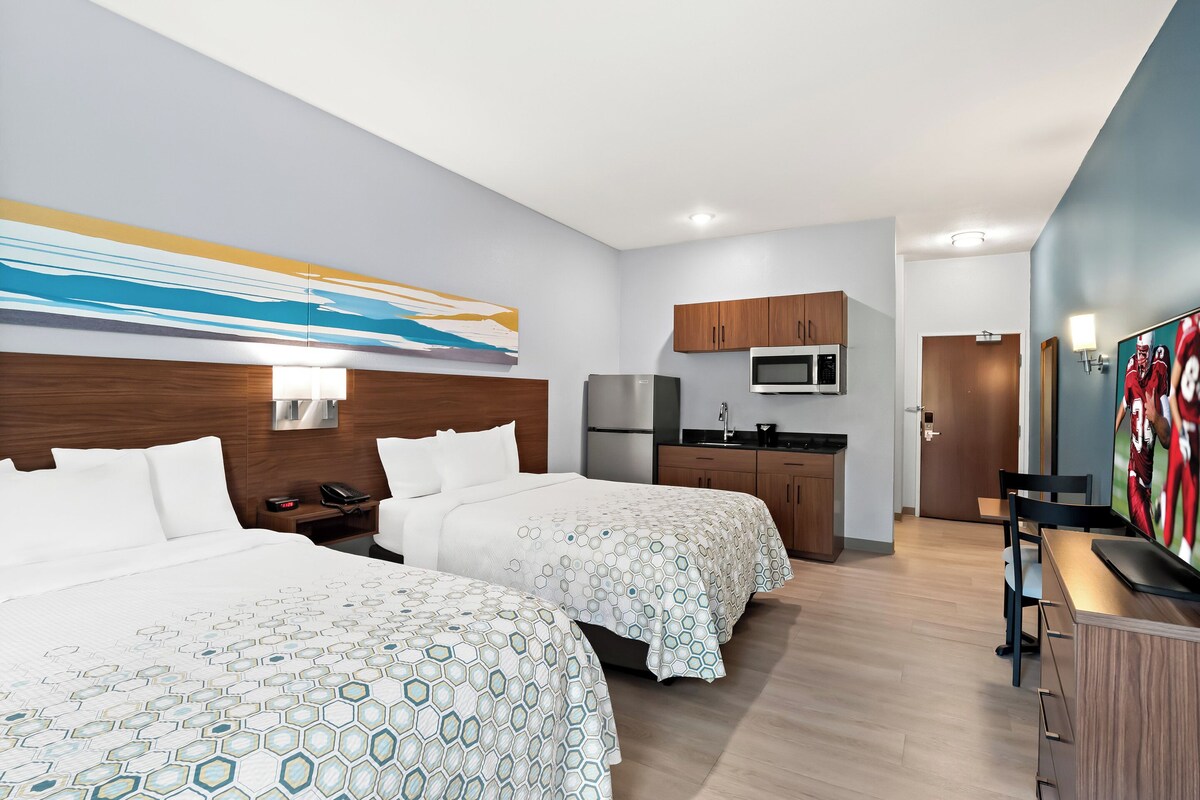 A hotel room with two beds, a kitchenette, a TV, and a painting on the wall.
