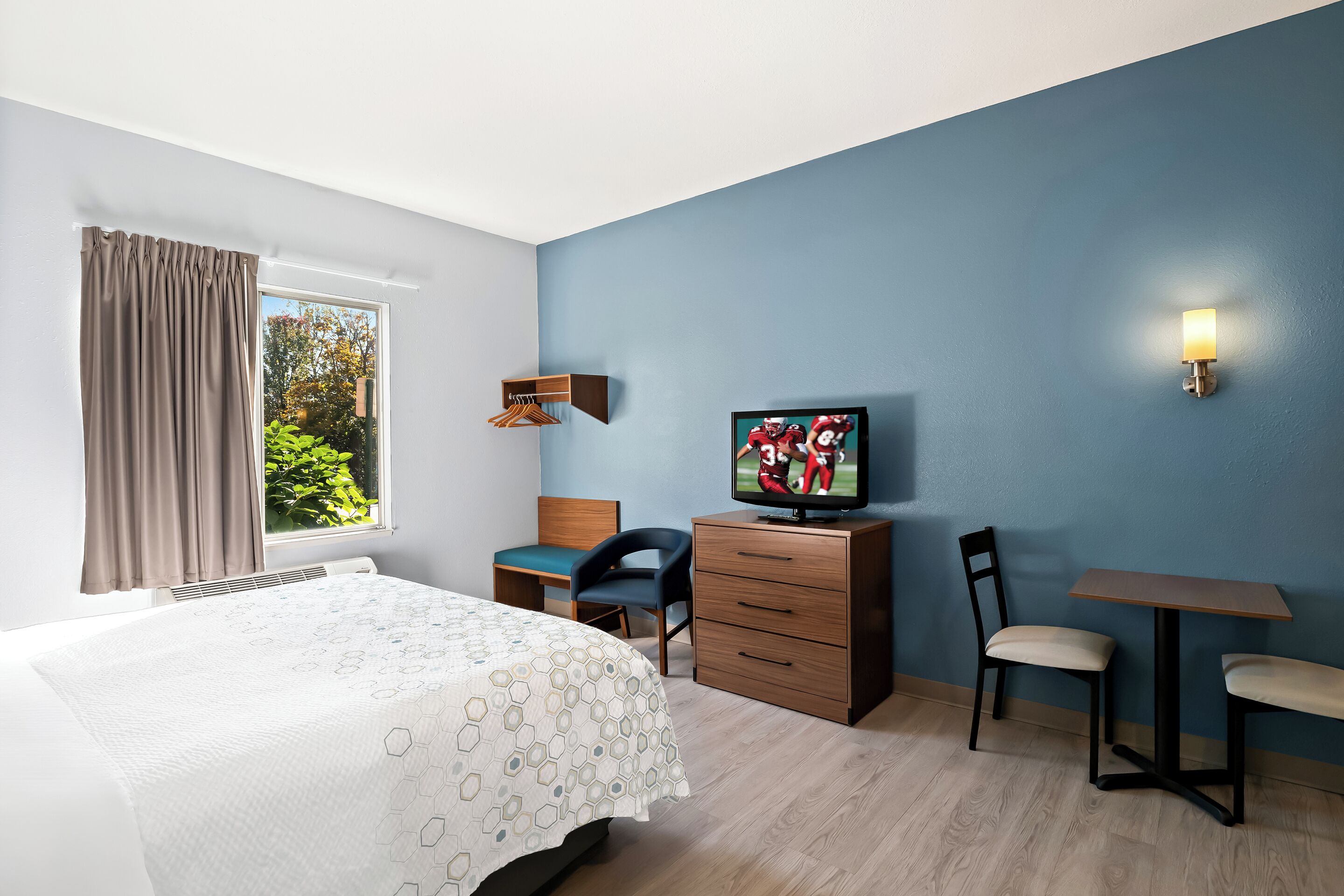 A hotel room with a bed, a desk, a chair, a TV, and a window with curtains.