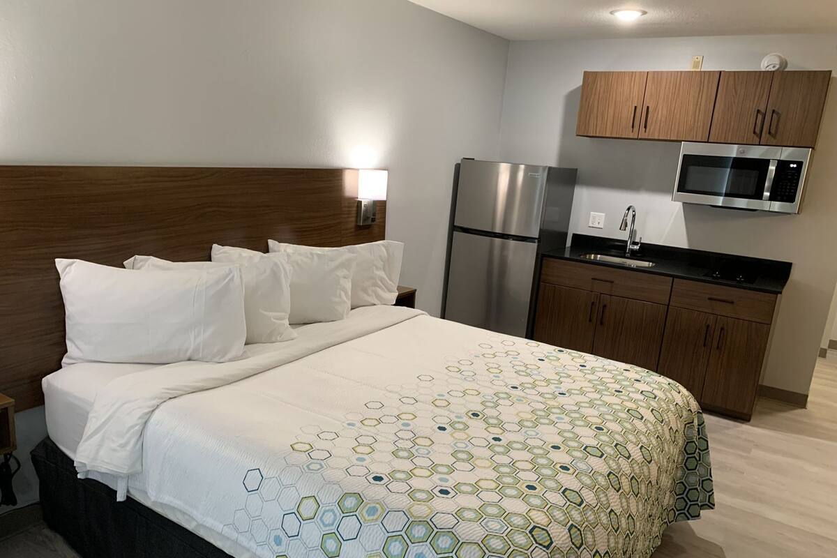 A hotel room with a bed, a refrigerator, a microwave, wooden cabinets, and a nightstand.