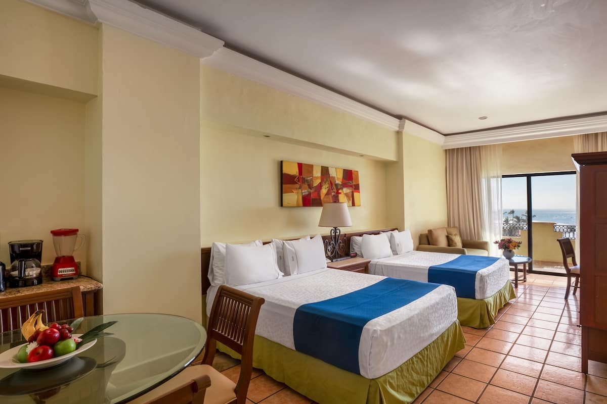 A hotel room with two beds, a dining table with fruit, a coffee maker, and a balcony with a view.