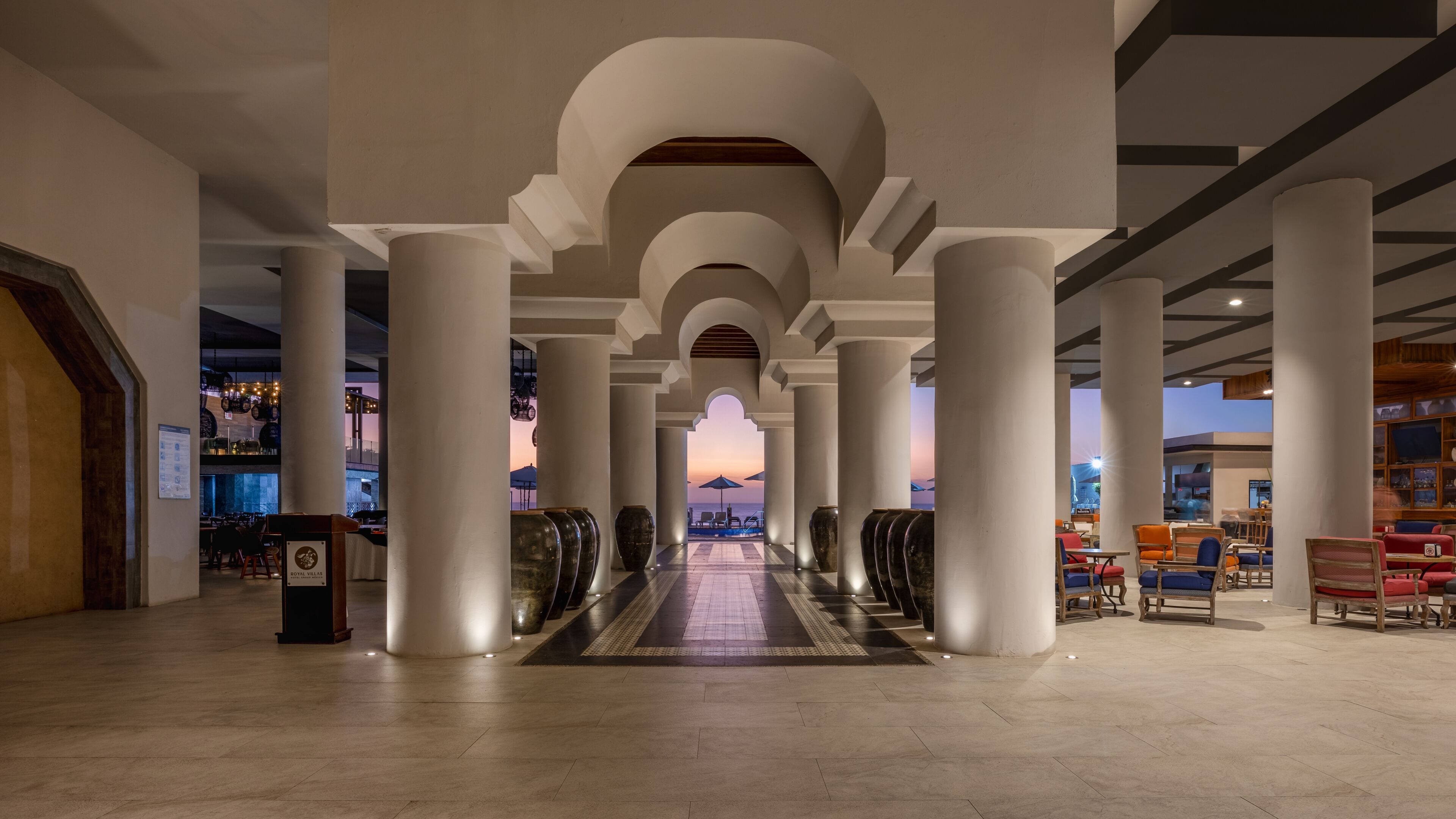 Pasillo de hotel con vista al mar al atardecer, que presenta un pasillo central flanqueado por columnas y zonas de estar.