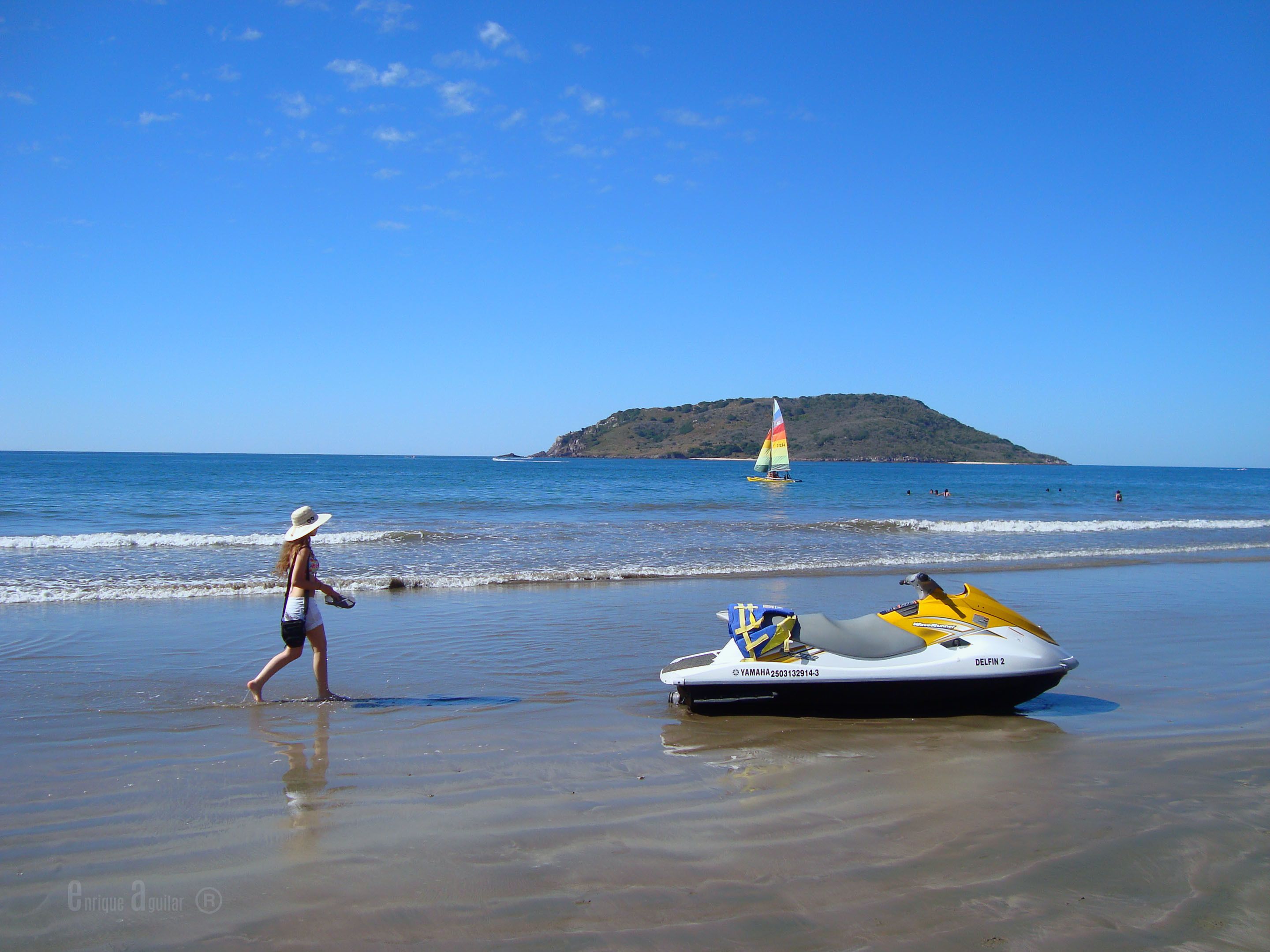 Une plage avec un jet ski et un voilier sur l’eau, une personne qui se promène sur le rivage et une île au loin.
