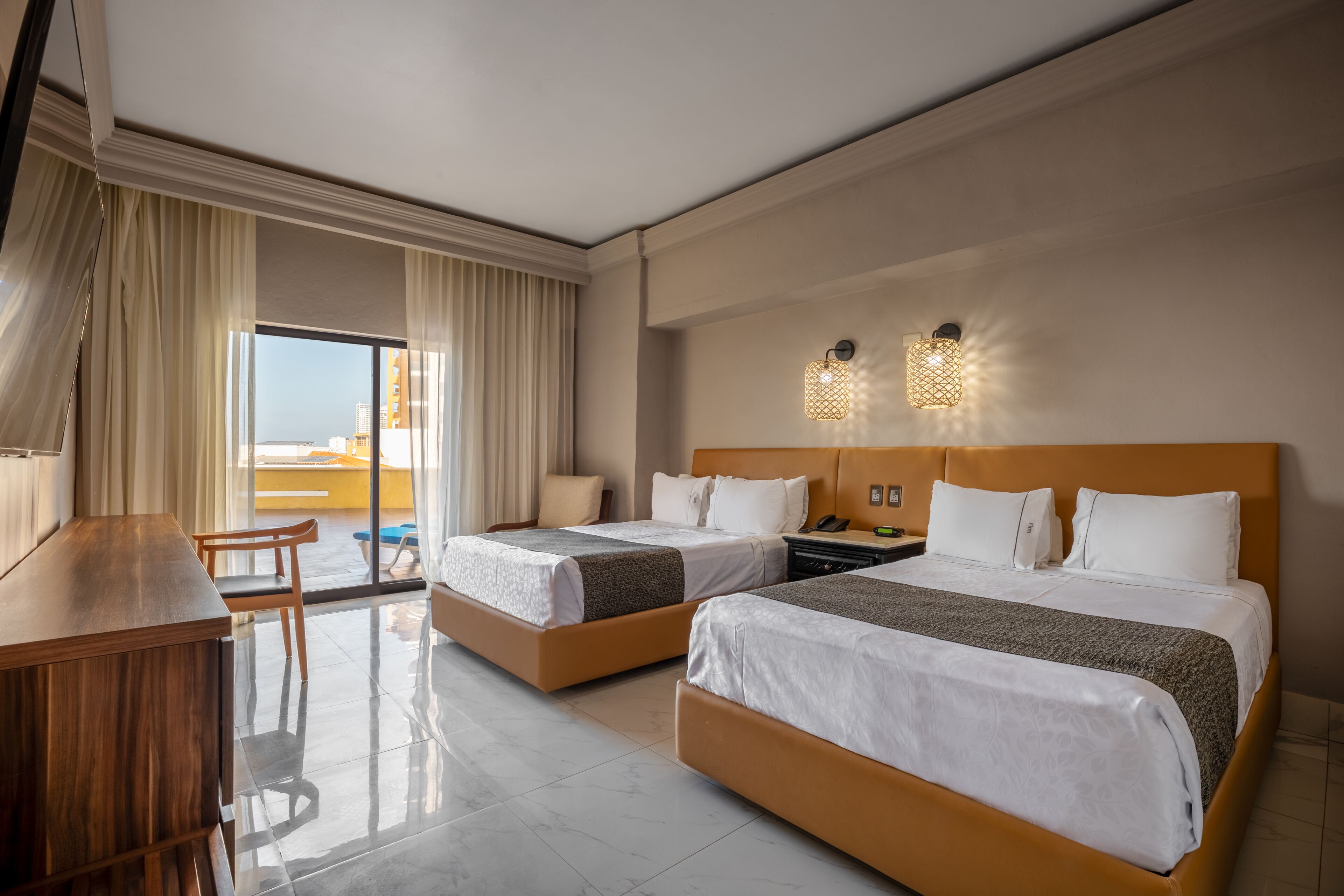 Habitación de hotel con dos camas, un escritorio, una silla, una mesita de noche y vistas a la ciudad.