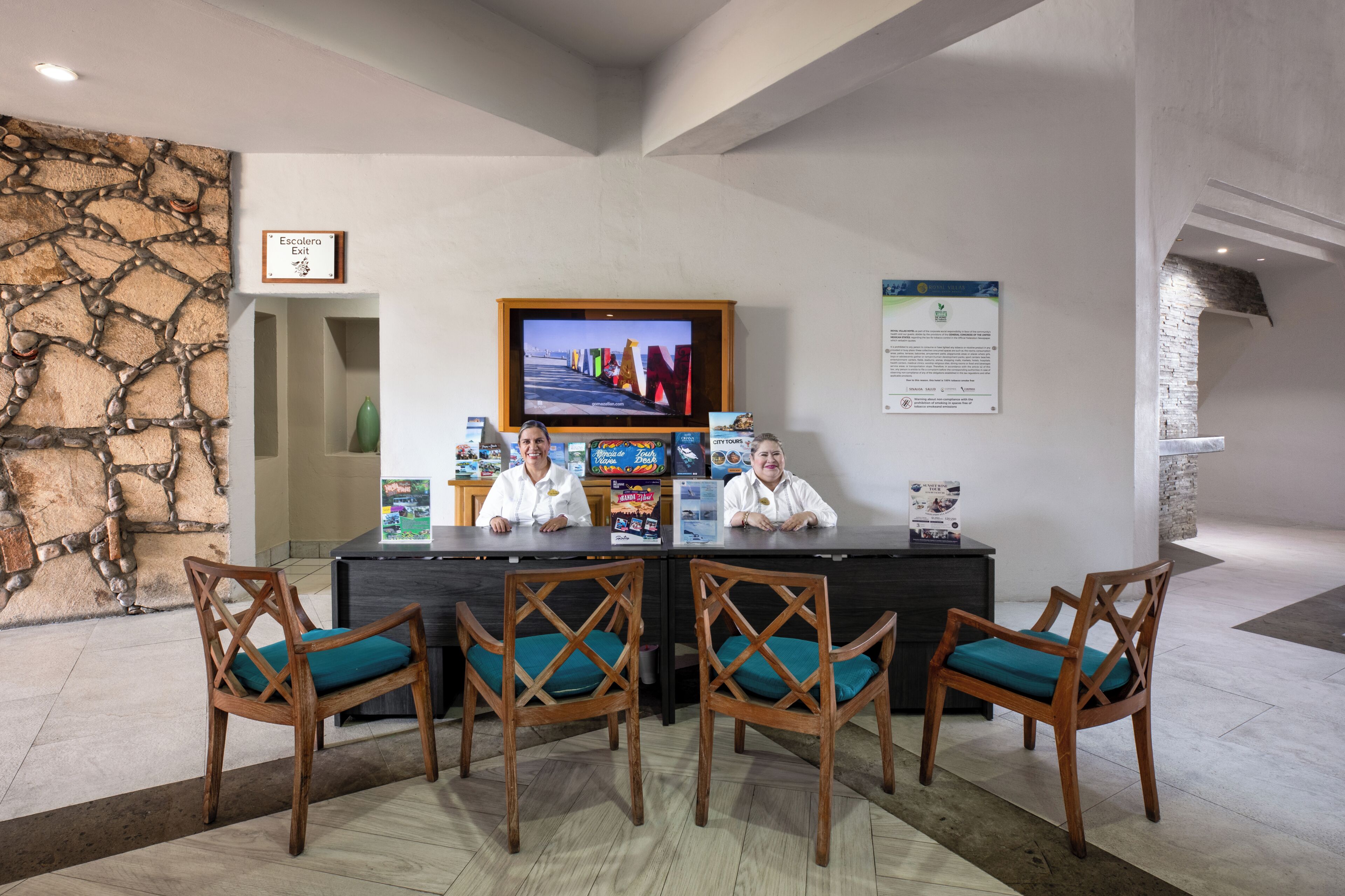 A reception desk with a television, framed exit sign, informational posters, and wooden chairs with teal cushions.