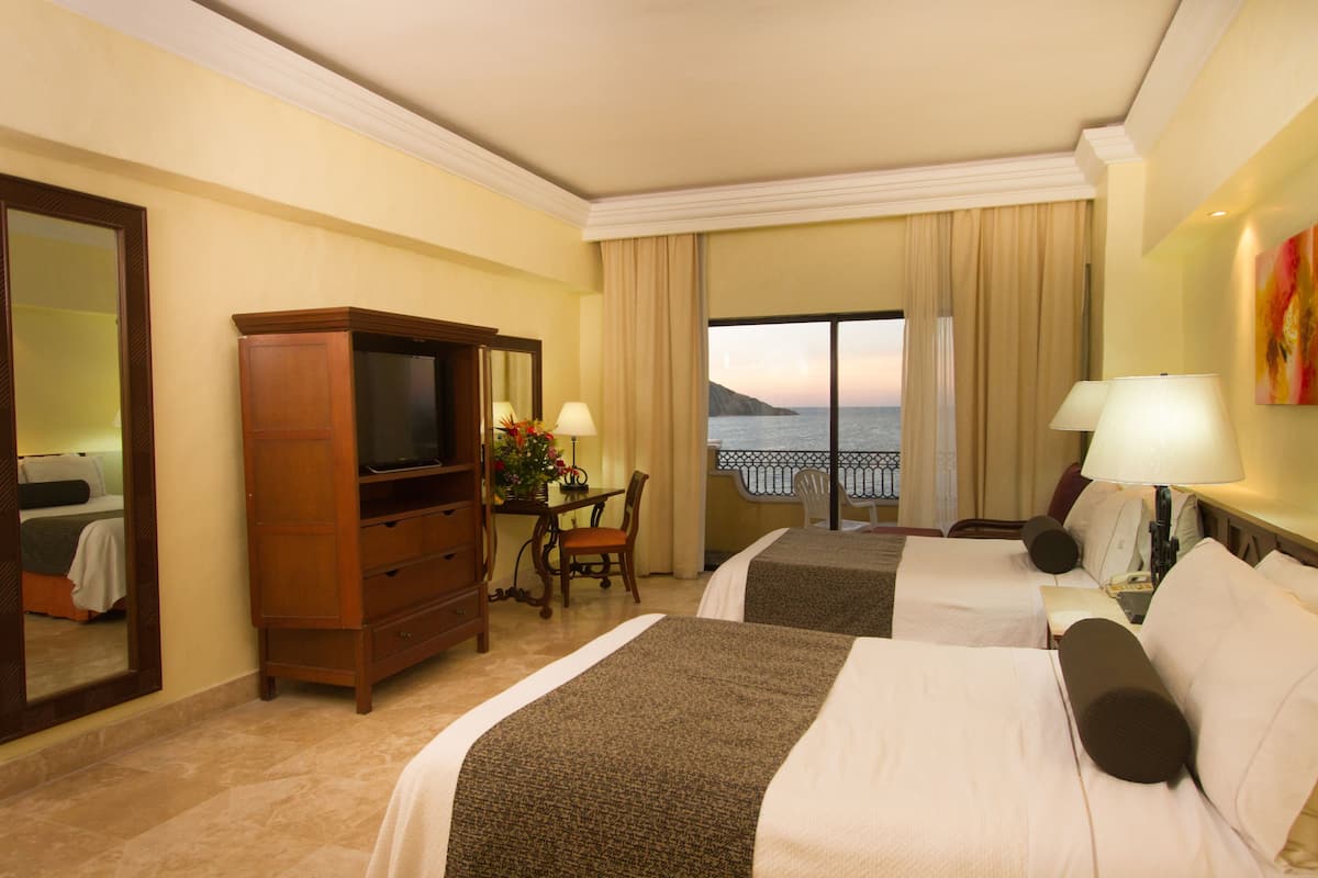 A hotel room with two beds, a desk, a television, and a view of the sea.