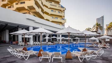 Un área junto a la piscina con sillones y sombrillas, una alberca y un edificio con balcones.
