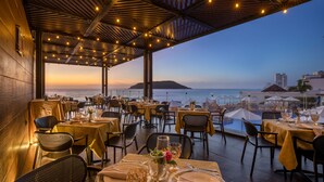 An outdoor dining area with tables set for two, overlooking a body of water and a distant island. - Royal Villas Resort (Mazatlán)