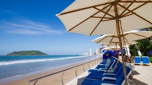 A beach with blue lounge chairs and white umbrellas, a sandy shore, and a view of the ocean and a distant island. - Royal Villas Resort (Mazatlán)