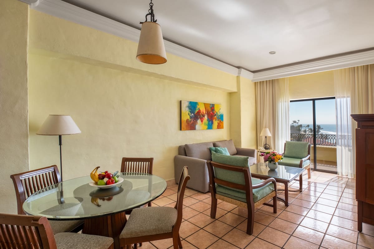 A dining area with a glass table, chairs, and a bowl of fruit. There is a sofa and a chair with a view of the ocean.