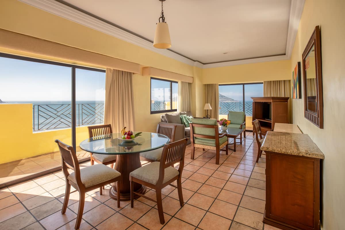 A hotel room with a balcony, a dining table with chairs, a sofa, and a TV stand.