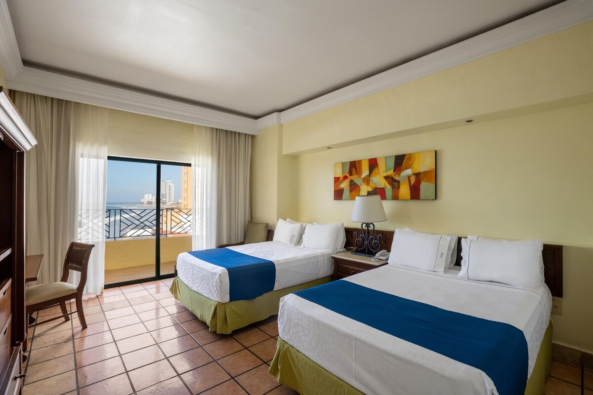 A hotel room with two beds, a balcony with curtains, a painting on the wall, and a view of the cityscape.