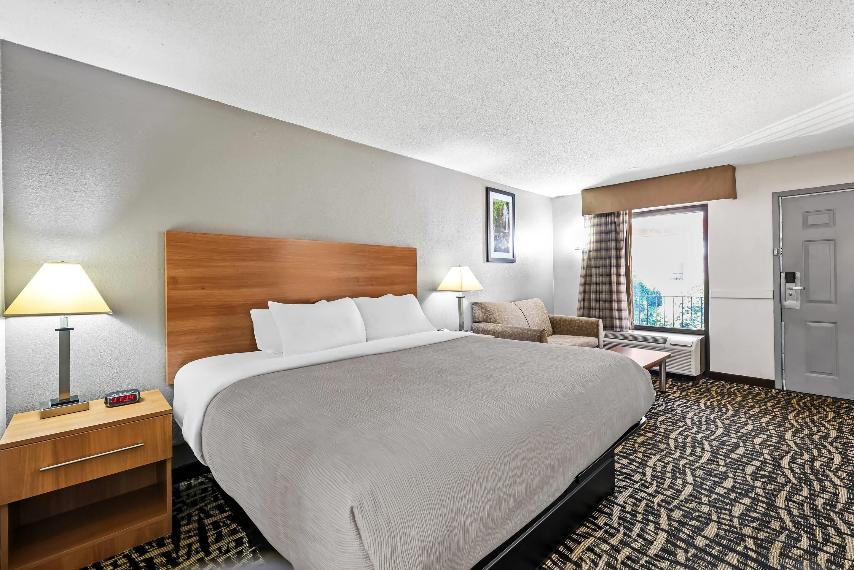Une chambre d’hôtel avec un grand lit, deux tables de chevet avec des lampes, une chaise, une fenêtre avec des rideaux et une porte.