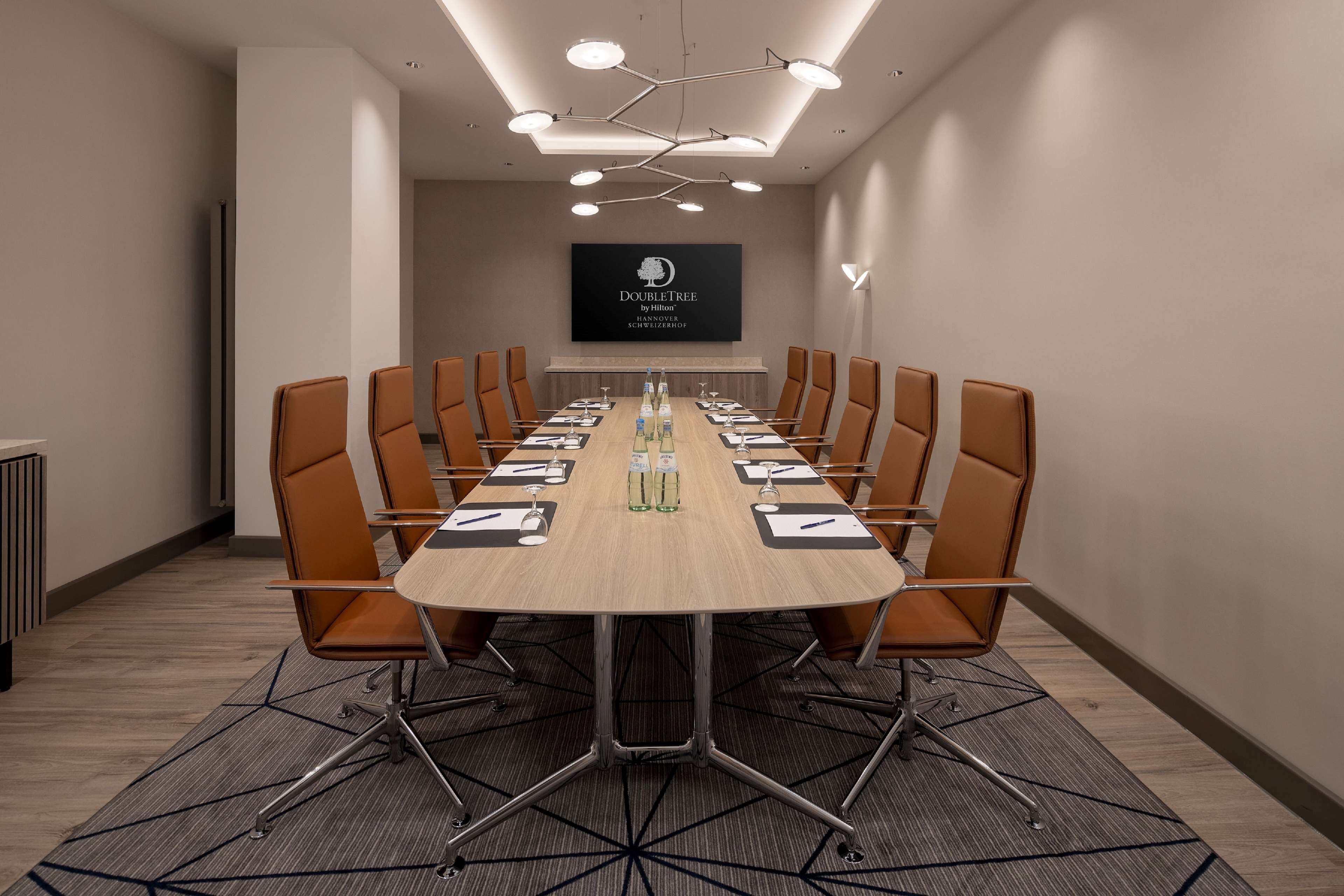 Une salle de conférence dotée d’une longue table, de fauteuils en cuir marron et d’un lustre moderne.