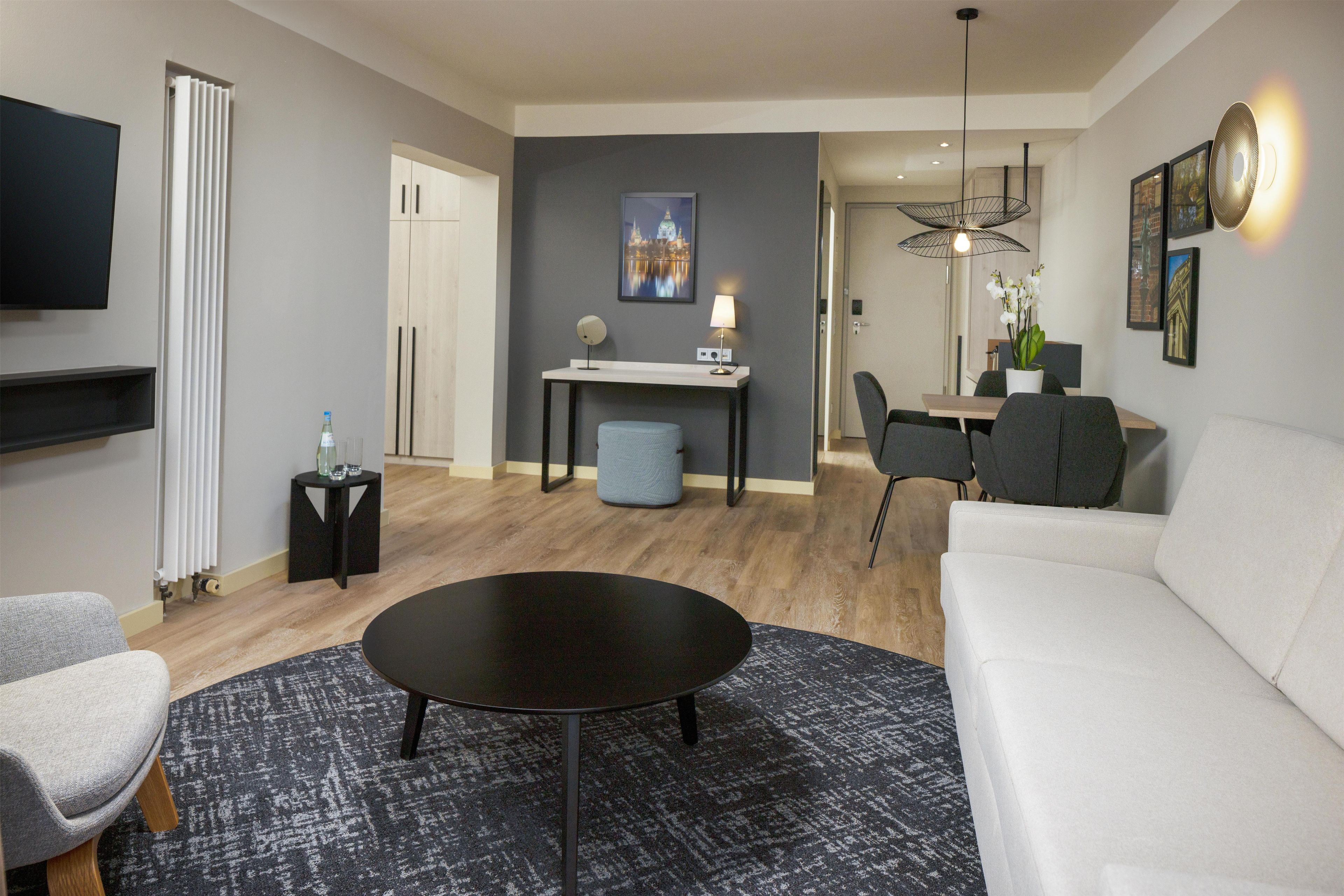 Uma sala de estar moderna com um sofá branco, uma mesa de centro preta e uma poltrona cinza. Há uma área de jantar com mesa e cadeiras, uma TV na parede e um radiador.