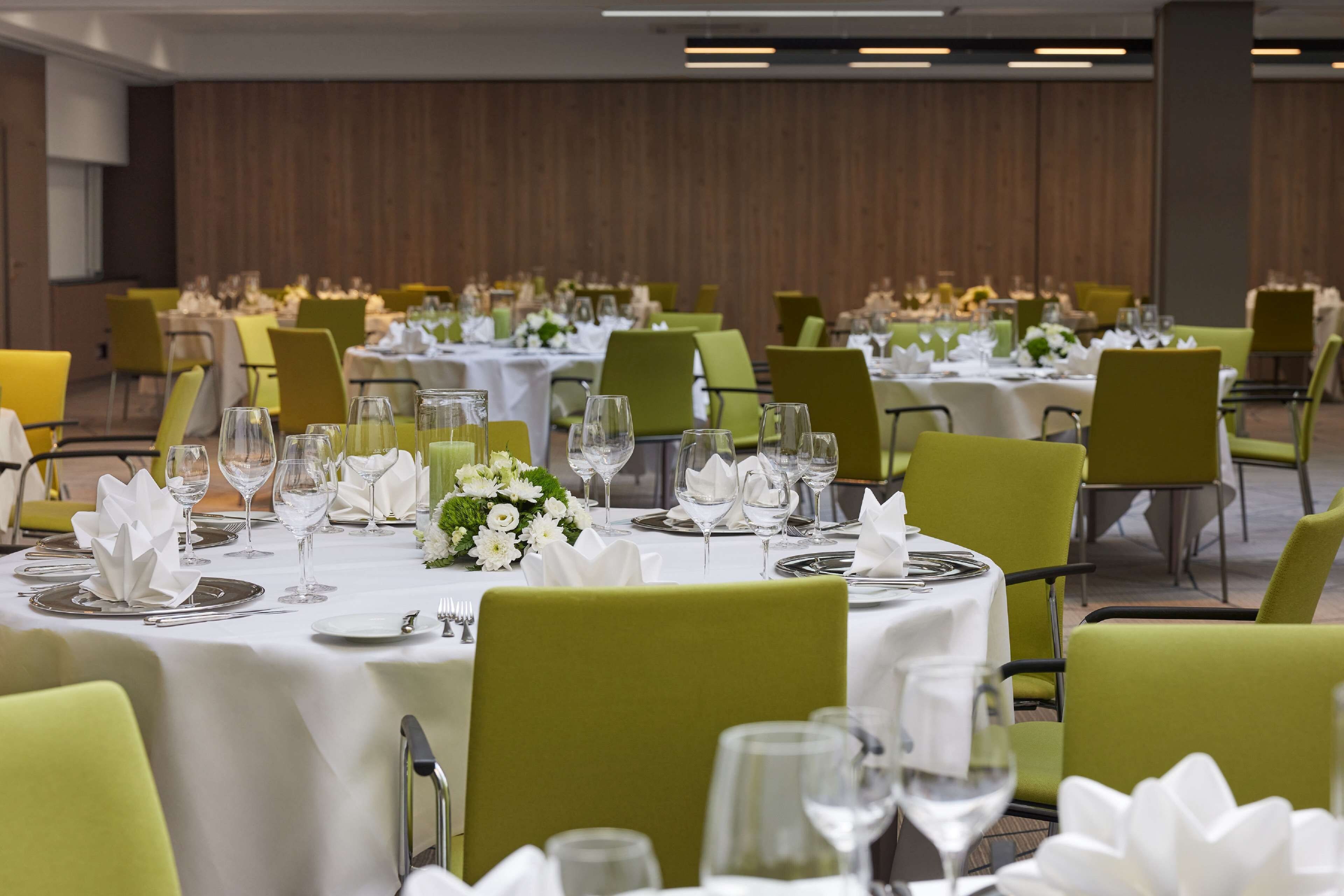 Salão de banquetes com mesas postas para refeições, cadeiras verdes e toalhas de mesa brancas.