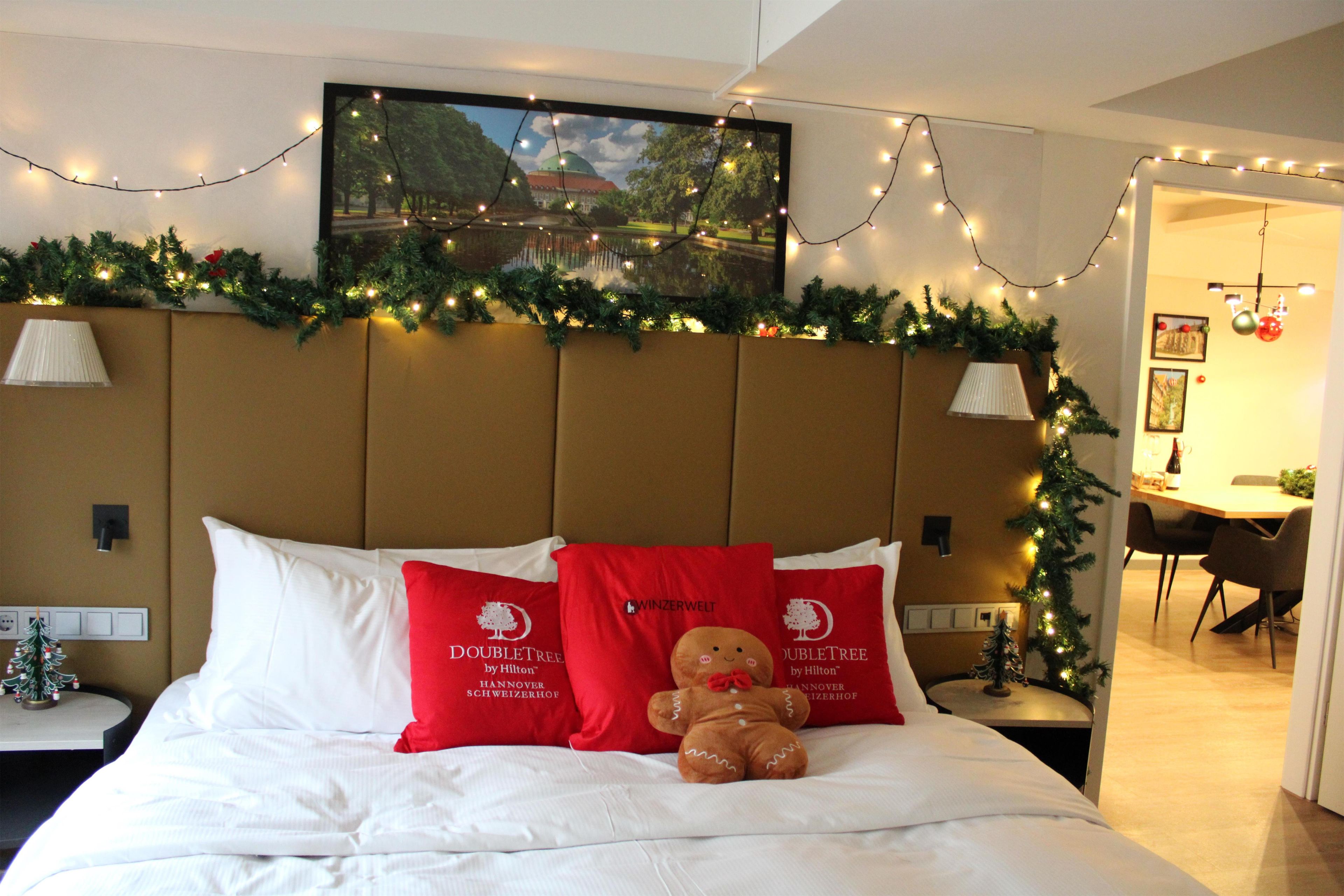 A hotel room with a bed, pillows, a teddy bear, a Christmas tree, and a painting on the wall.