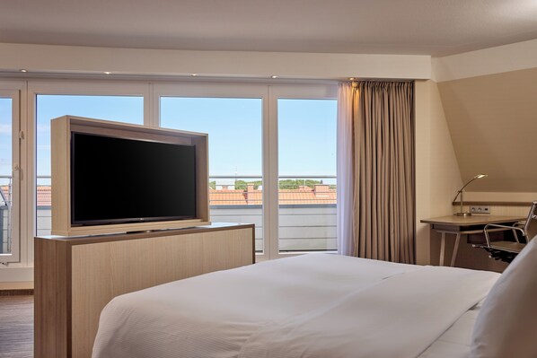 Habitación de hotel con una cama grande, un televisor de pantalla plana sobre un soporte de madera, un escritorio con lámpara y vistas a la ciudad a través de amplios ventanales.