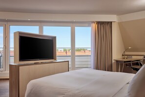Habitación de hotel con una cama grande, un televisor de pantalla plana sobre un soporte de madera, un escritorio con lámpara y vistas a la ciudad a través de amplios ventanales.