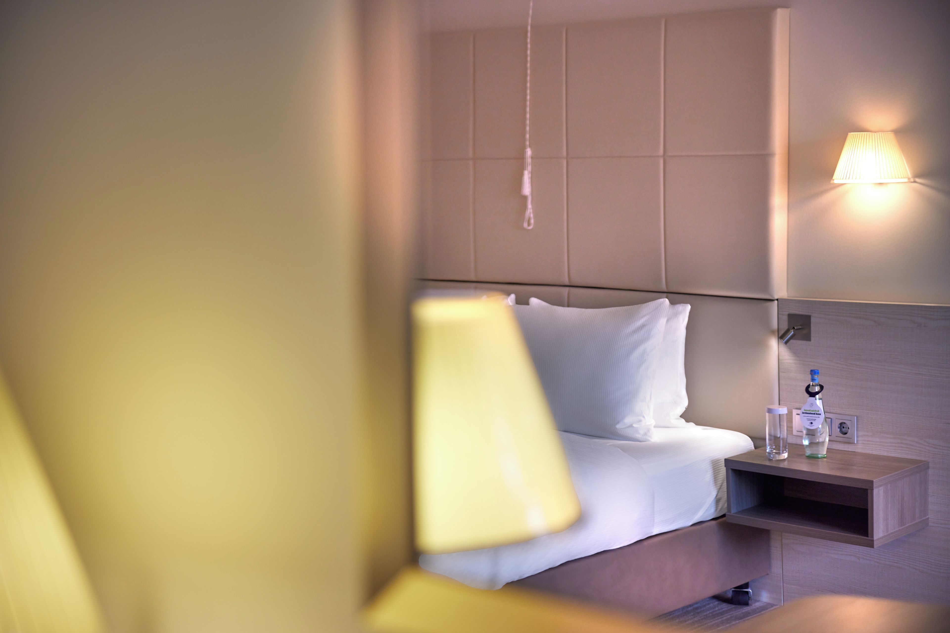Una habitación de hotel con cama, mesita de noche, lámpara y luz en la pared.