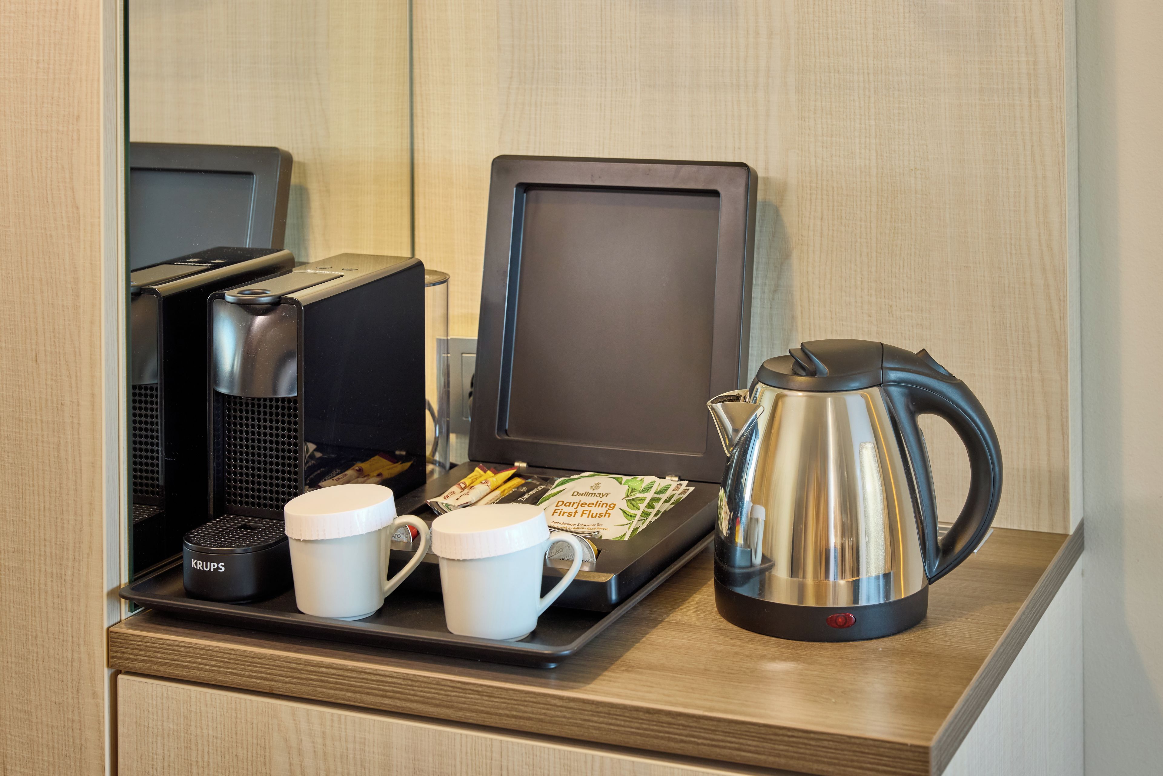 Una estación de café con una cafetera, dos tazas, una bandeja con sobres de café y un hervidor.