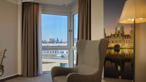 Habitación de hotel con un ventanal grande, una silla, una lámpara y un mural en la pared que representa un edificio.