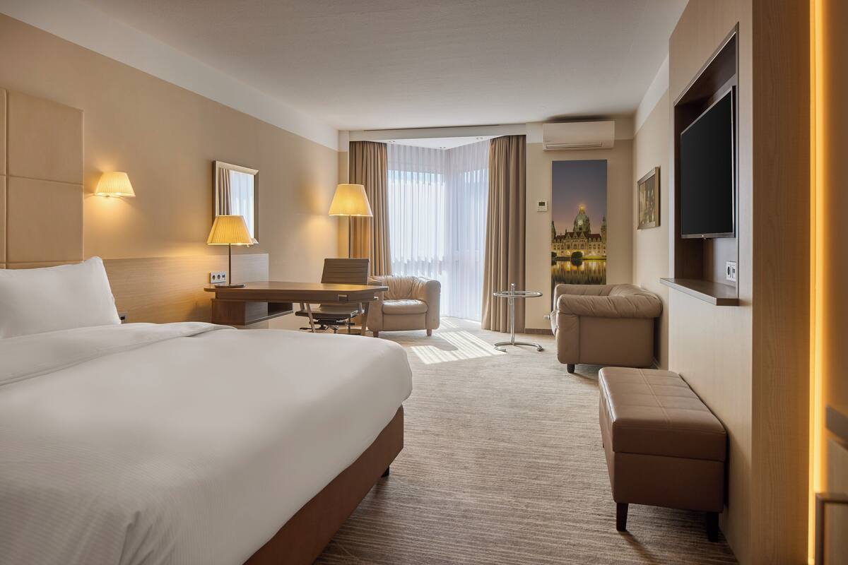 A hotel room with a large bed, a desk, two chairs, a small table, a sofa, and a TV.